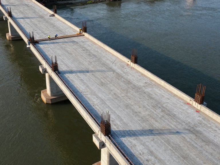 Aerial view of a concrete bridge deck under construction spanning a waterway, reinforcement rebar visible in sections of formwork, construction crew working in the far span, late afternoon light casting long shadows across the deck surface