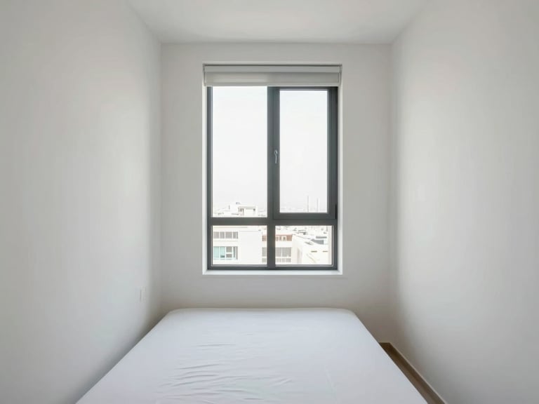 Full-room shot of a compact Dubai apartment bedroom, clean white walls, minimal furniture, even window daylight revealing proportion and finish quality, no people