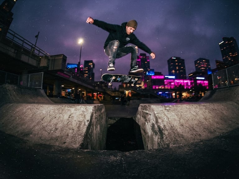 Dynamic low-angle wide shot of a skateboarder caught mid-air over a concrete gap at dusk, illuminated by vibrant magenta and blue streetlights, deep purple overlay, gritty 35mm film texture.