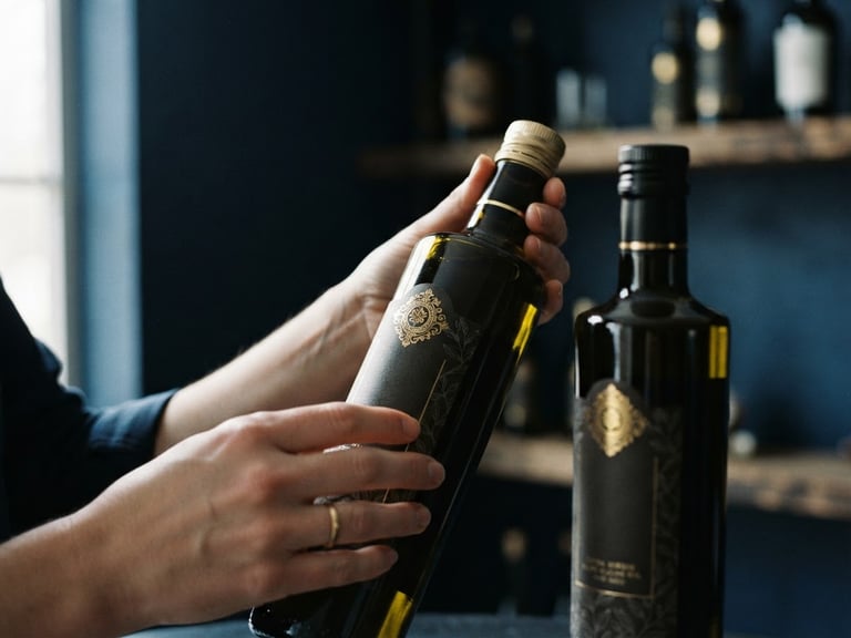 Moody editorial food photography, close-up of premium olive oil bottles being carefully inspected under soft natural side-light, gold accents, deep navy background.