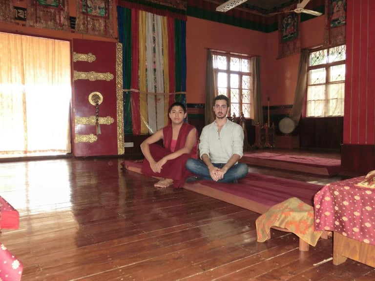 A Buddhist monk and a traveler sit cross-legged on mats inside a traditional Tibetan temple sanctuary.