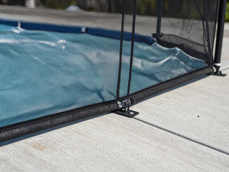 Close-up environmental shot, a section of removable mesh pool fence anchored into a concrete deck, natural daylight from above, camera at child's eye level showing the fine mesh weave and the tamper-resistant sleeve anchor flush with the deck surface