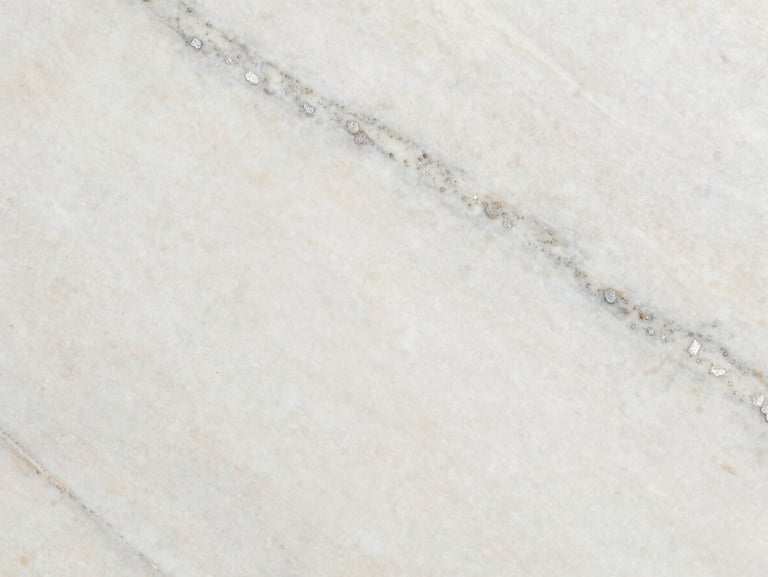 Macro shot of Pearl White granite slab surface under controlled studio raking light — creamy off-white ground with soft grey mineral streaks and faint silver mica flecks, honed finish showing subtle surface texture without high gloss