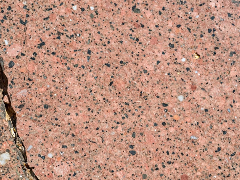 Close-up of Cheema Pink granite slab under direct natural daylight — warm rose-pink feldspar ground with dark biotite clusters and white quartz pockets, polished finish, raking light from the left edge revealing surface crystalline detail
