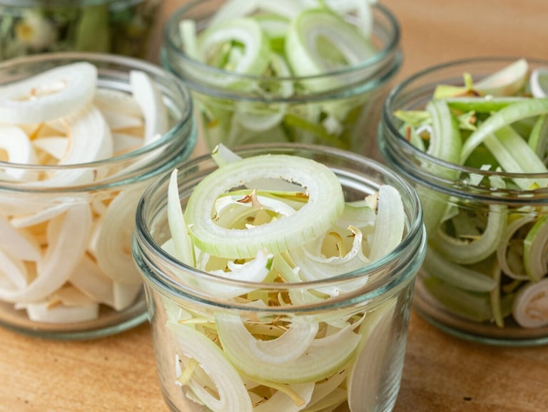 Close-up of dehydrated sliced onions and garlic flakes in clean industrial glass containers on a wooden table, bright daylight, sharp focus.