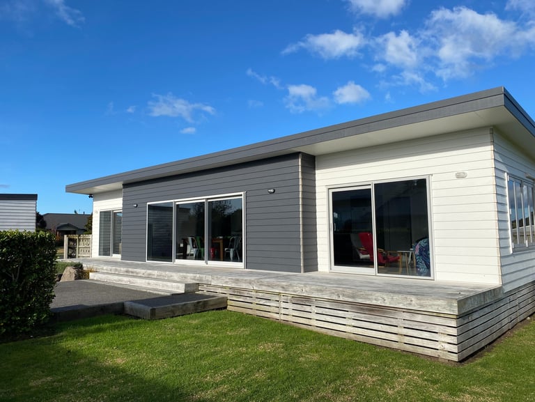 Modern prefabricated home featuring charcoal and white siding with a large wooden deck and glass sliding doors.