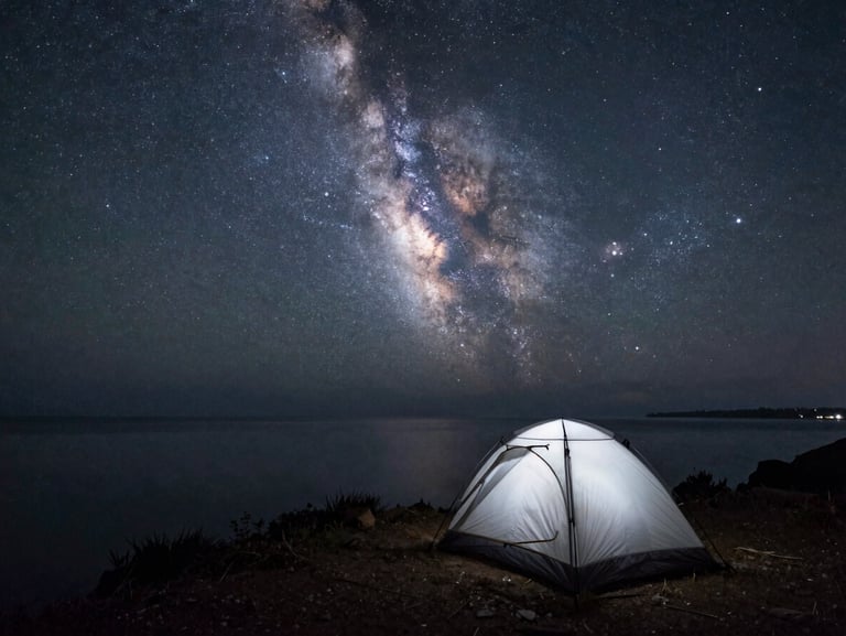 Wide starscape over a single silhouetted tent pitched on a coastal cliff, the Milky Way glowing softly above a calm dark sea.