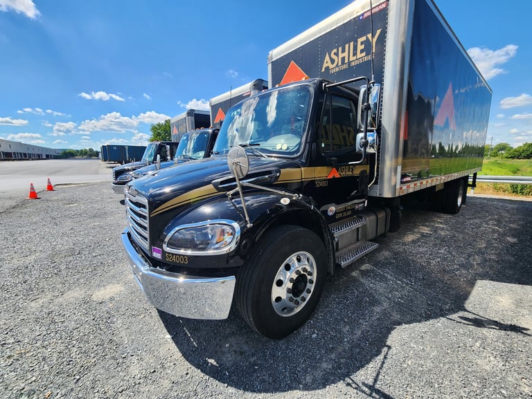 Ashley Furniture delivery trucks parked in a gravel lot on a sunny day.
