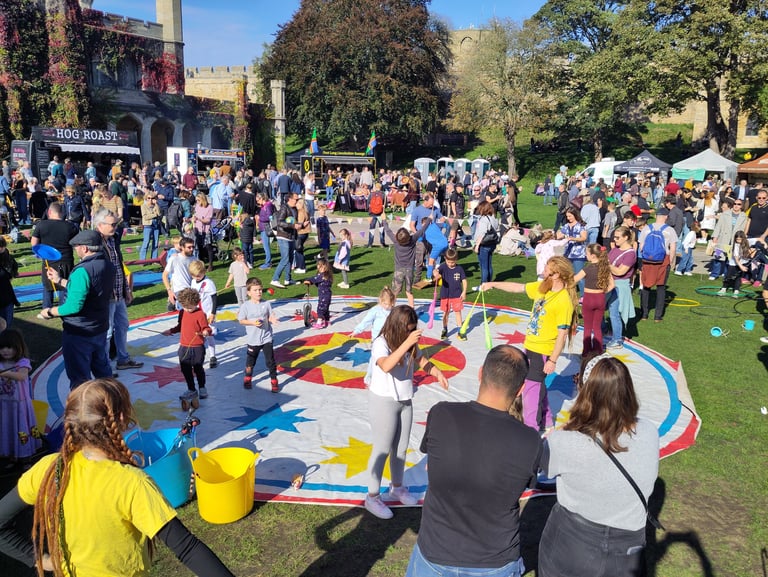 Crowds of people and children enjoying a sunny outdoor festival with circus skills and food stalls.
