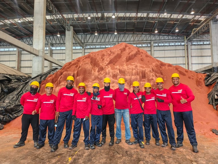 Industrial warehouse workers in red uniforms and yellow hard hats standing before a large pile of mineral ore.