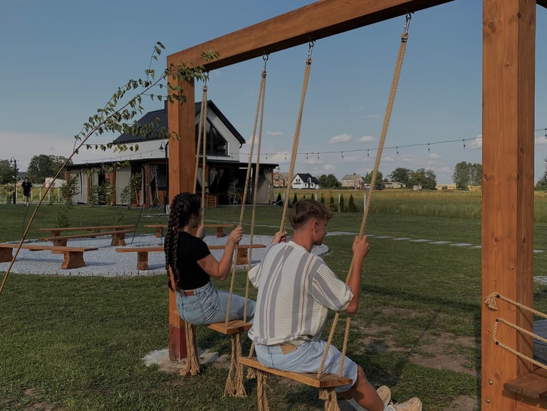 Dwie osoby na drewnianej huśtawce, a w tle domek