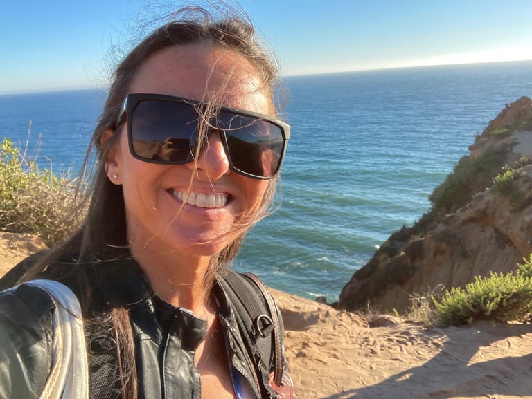 Michael Alexa Pitts smiling in the sunshine wearing black shades, a rocky Pacific coastline, blue sky & blue ocean behind
