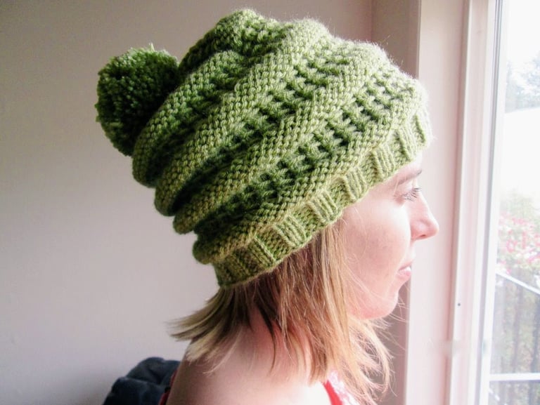 A young woman staring out a window wearing a handknit green slouchy beanie with a pompom