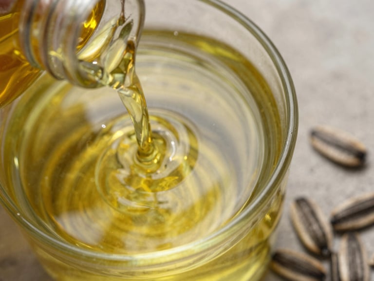 Extreme close-up overhead of amber sunflower oil poured into a clear glass vessel on a stone surface, natural daylight, oil surface reflecting light, pressed sunflower kernels scattered nearby