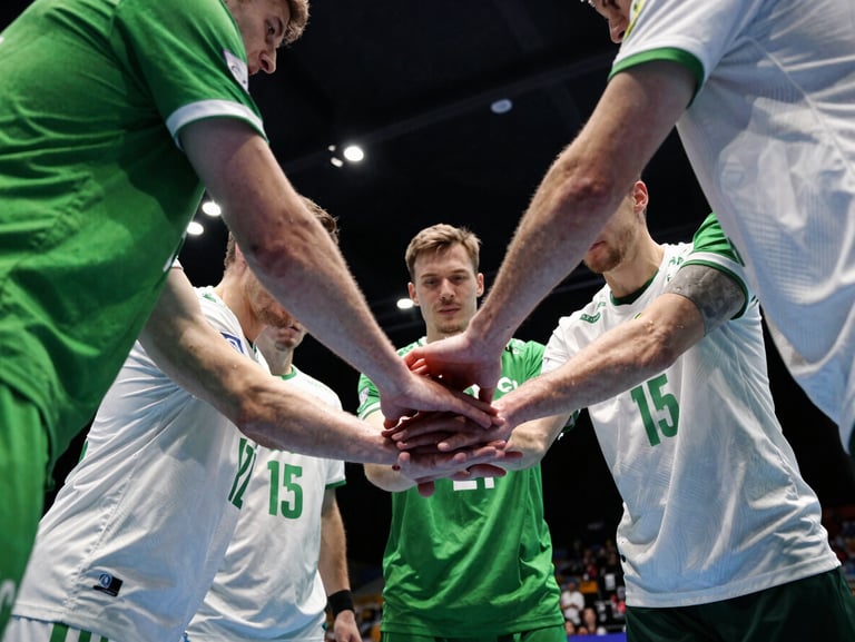 Intense team huddle of handball players in green and white uniforms, hands stacked in the center, dramatic sports hall lighting, high-contrast
