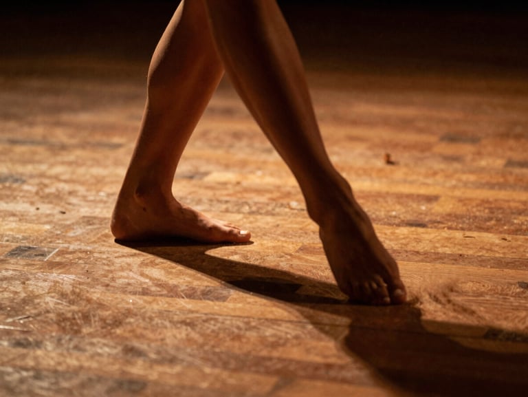 Close-up of a dancer's feet in low-angle light, dust kicked up from the wooden stage floor, high-contrast shadows, warm editorial style.
