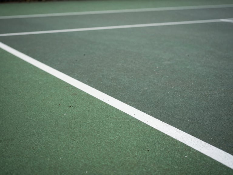 Close-up of a weathered green macadam tennis court surface, white painted baseline, soft natural overcast UK daylight.