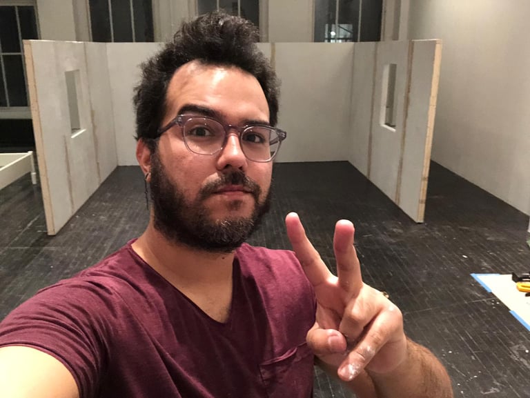 A man with glasses and a beard making a peace sign in an art studio with white partitions.