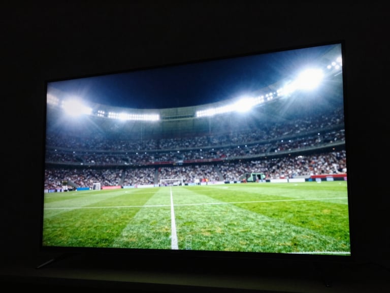 Low-angle high-contrast shot of a glowing smart TV screen in a pitch-black room, displaying a vibrant green football pitch under bright floodlights, dramatic backlighting.