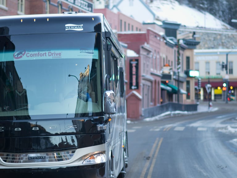 A luxury Newmar Essex motorhome driving through a snowy mountain town street with historic buildings.