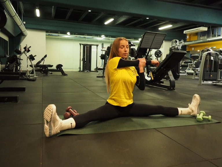 Una donna in maglia gialla esegue uno stretching in spaccata seduta, allungando il braccio per un esercizio di mobilità,