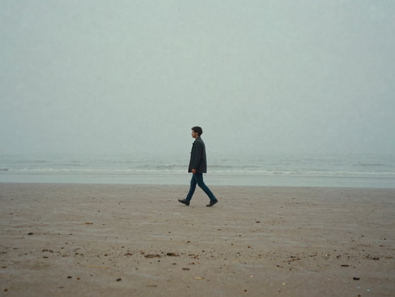 Cinematic 35mm still of a lone figure walking on a foggy beach, muted blue and gray tones, wide landscape framing, minimalist composition.