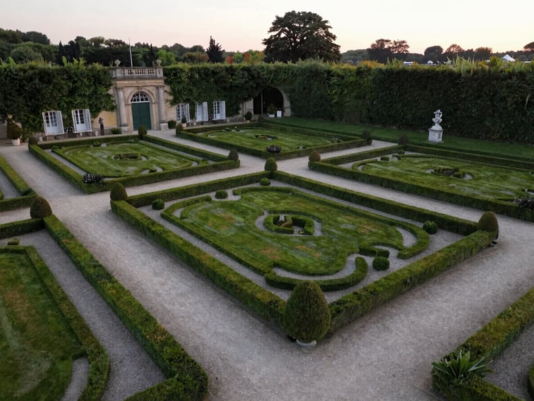 An aerial wide shot of a classic French estate garden with perfectly manicured hedges and gravel pathways, soft evening light.