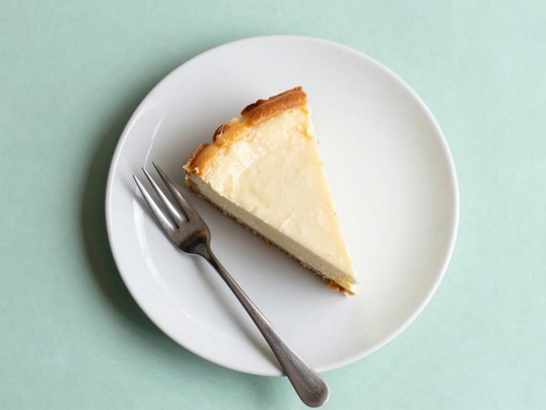 Overhead flat-lay of a single slice of creamy cheesecake with a golden crust on a matte white ceramic plate, a delicate silver fork beside it, soft natural daylight, mint green background, 35mm