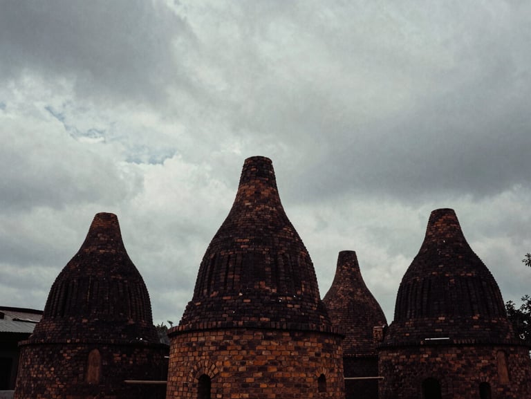 Atmospheric, high-contrast 35mm photograph of brick kilns in the distance under a heavy, cloudy sky, desaturated tones, deep shadows.