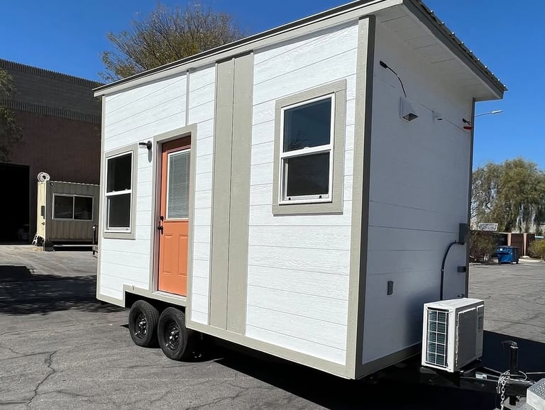 A white tiny house on wheels with a bright orange door and a mini-split AC unit parked on asphalt.