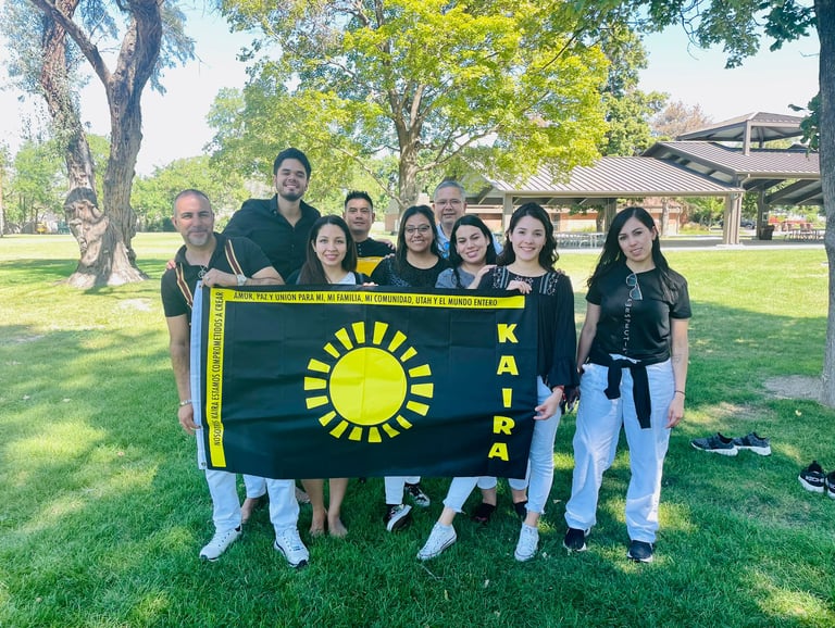 A diverse group of people posing in a park holding a black KAIRA flag with a sun logo.