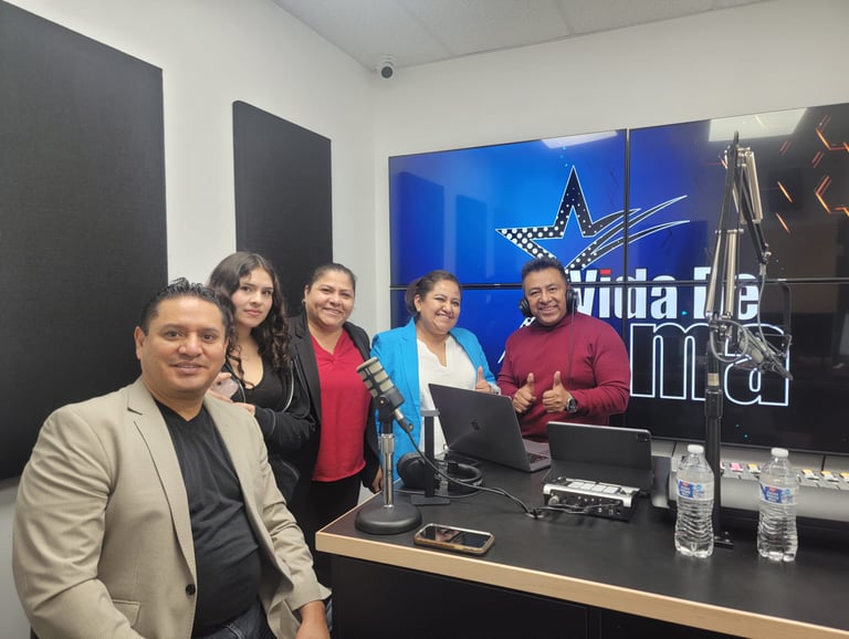 A group of five smiling professionals posing in a modern radio broadcasting studio with microphones and digital displays.