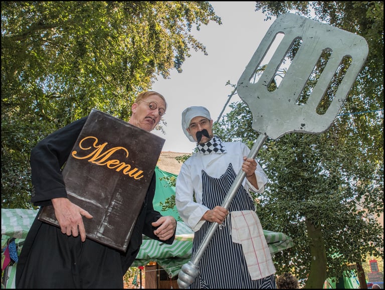 Two street performers in chef costumes holding a giant menu and an oversized metal spatula outdoors.