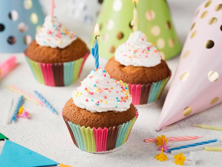 Birthday cupcakes with white frosting, sprinkles, and a lit candle next to party hats.