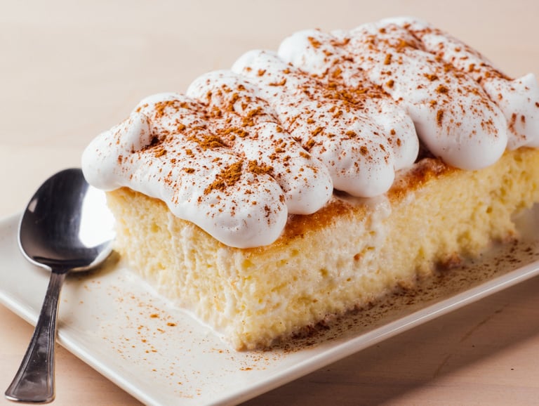 A slice of tres leches cake topped with whipped cream and cinnamon on a white plate with a spoon.