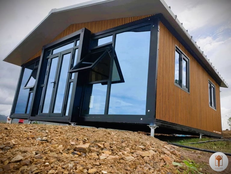 A modern prefabricated tiny home with wood siding and large glass windows on a rocky foundation.