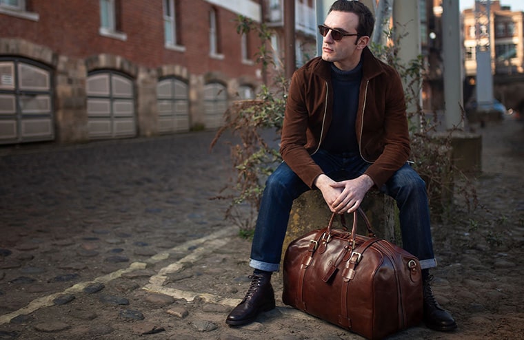 A stylish man in a brown jacket and sunglasses poses with a leather travel duffel bag on a cobblestone street.