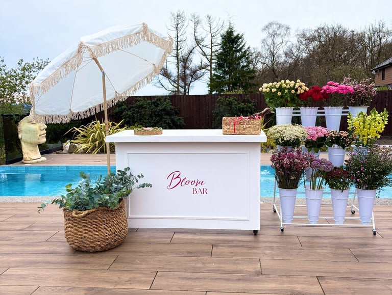 Mobile bloom bar flower cart with white fringed umbrella and fresh floral arrangements by a poolside deck.