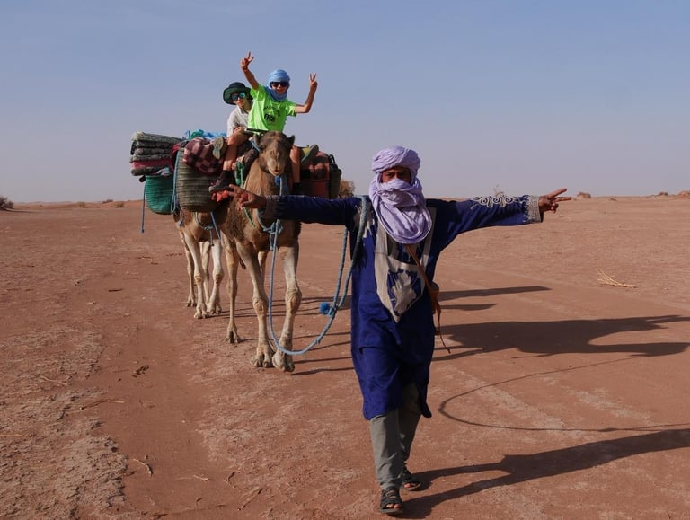 A desert guide in traditional dress leads two camels in the Sahara carrying children