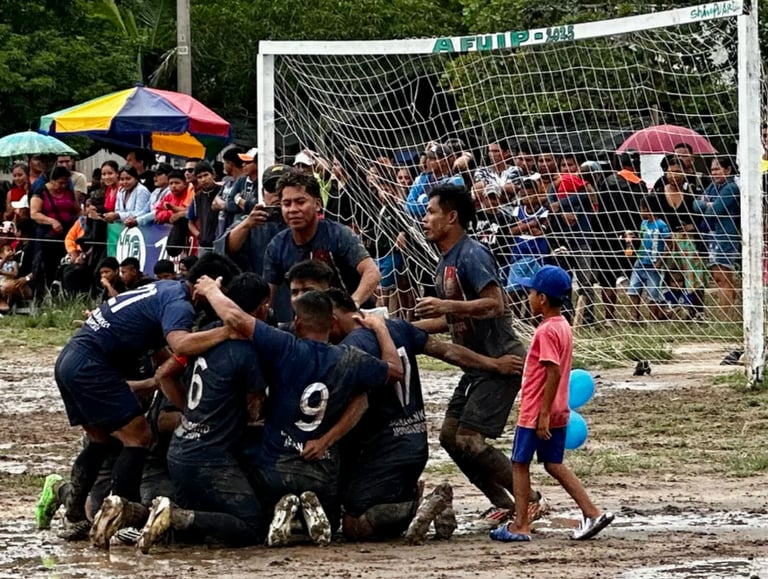 Muddy soccer players in blue jerseys celebrate a goal on a dirt field during a community football match.