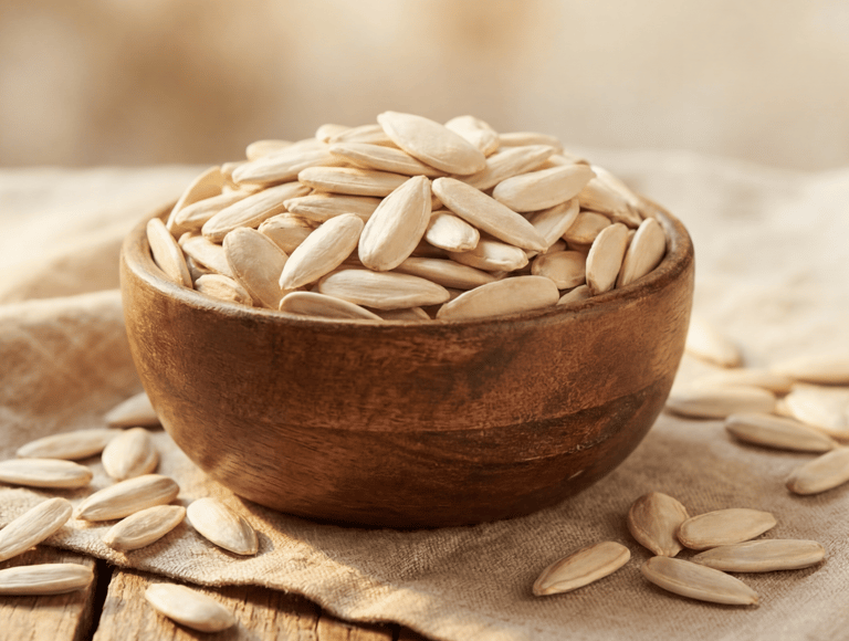 A rustic wooden bowl filled with roasted white pumpkin seeds on a burlap cloth.