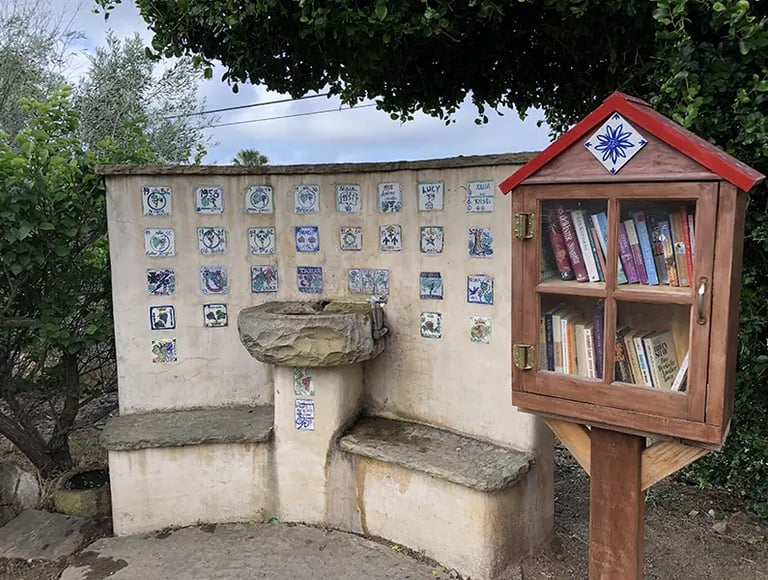 A stone drinking fountain with decorative tiles next to a wooden Little Free Library box filled with books.