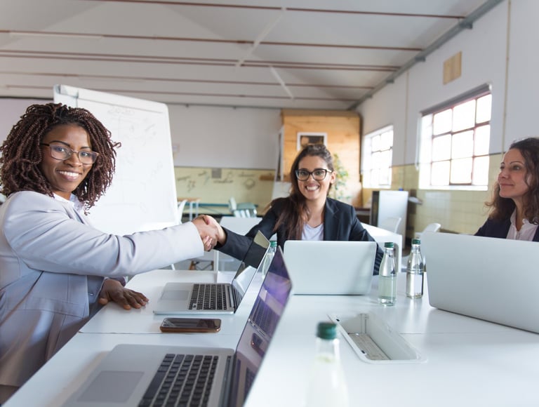 Professional business women in a modern office shaking hands over laptops during a successful meeting.