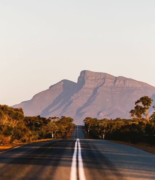 Empty asphalt road leading toward a massive mountain range during a golden hour sunset.