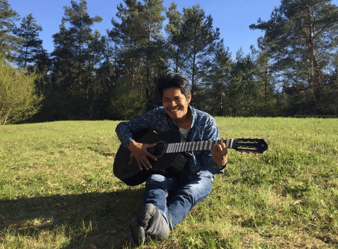 Roger Bardales playing an acoustic guitar while sitting on grass in a sunny park in Lithuania.