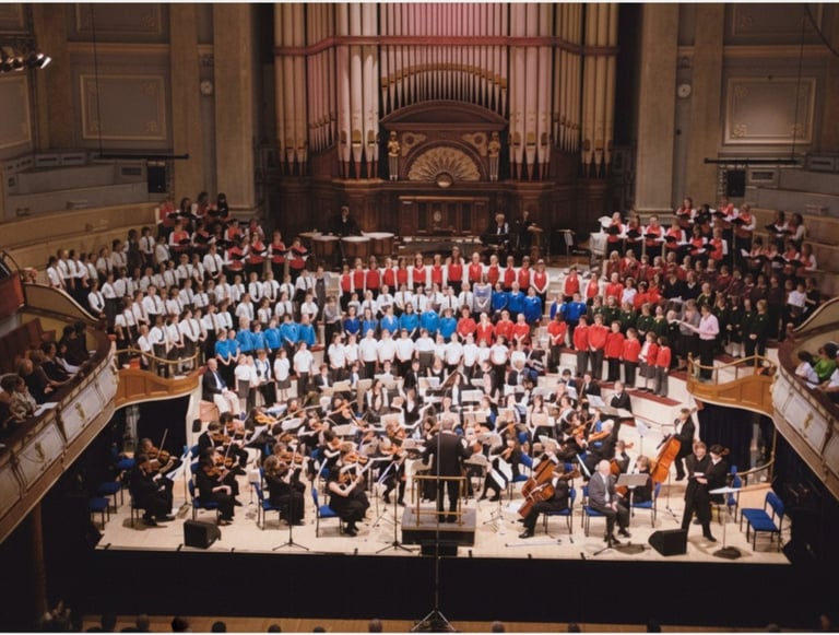 A large youth choir and symphony orchestra perform a concert on stage in Huddersfield Town Hall.