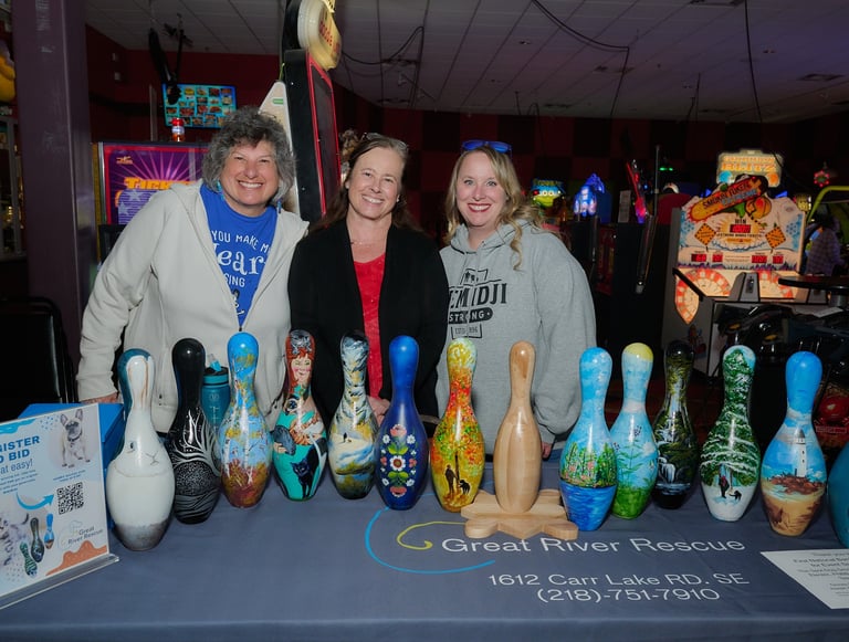 Three women smiling behind a display of hand-painted bowling pins for a Great River Rescue charity event.