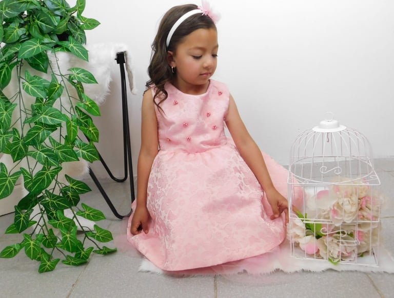 Niña con vestido de fiesta floral en tonos rosa, sentada junto a una jaula blanca decorada