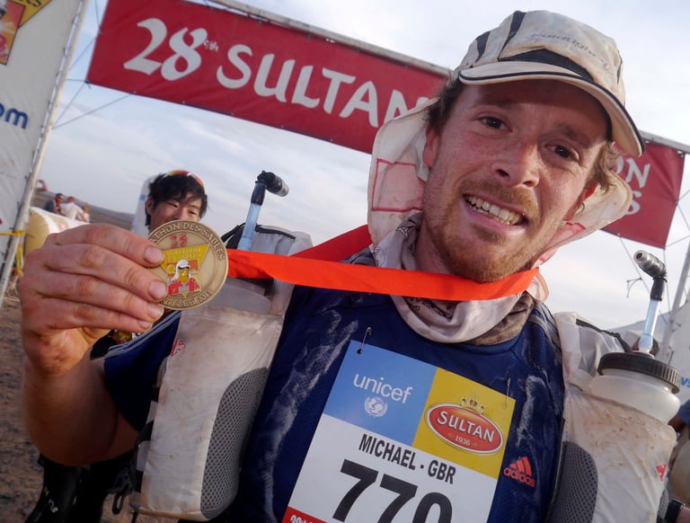 Smiling ultramarathon runner holding a gold medal at the Marathon des Sables finish line banner.