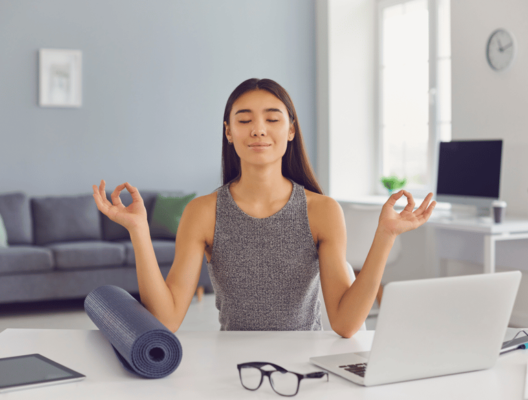 A female professional meditating in front her desk at home, featuring a computer and a yoga mat.